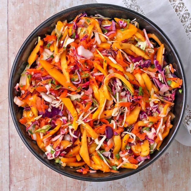 overhead shot of slaw with red and green cabbage, grated carrots, chopped cilantro and mango slices in a black bowl.