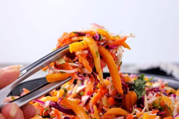 Silver tongs hold vegan coleslaw made with green and red cabbage, grated carrots, mango slices, and chopped cilantro.
