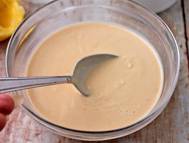 A glass bowl filled with lemon tahini dressing is stirred with a spoon.