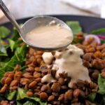 Lemon tahini salad dressing is ladled over a green salad with cooked lentils, red onions, and tomatoes.