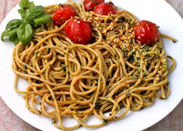A white pasta plate with spaghetti mixed with basil and spinach pesto, blanched tomatoes, hemp seed Parmesan, and fresh basil leaves.