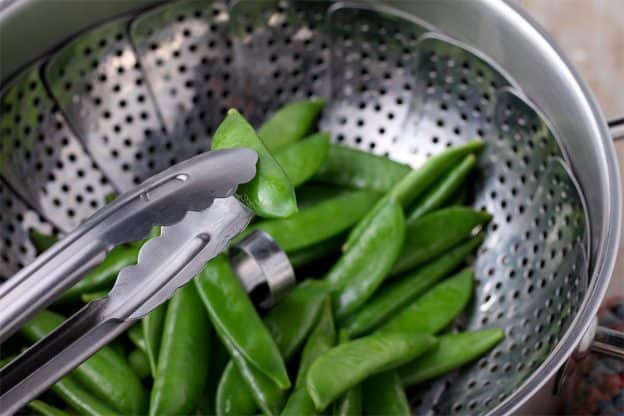Sugar snap peas are placed in a steamer basket with one pod held with silver tongs.