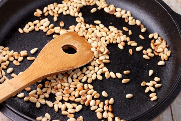 Pine nuts are toasted in a black skillet with a wooden spoon.