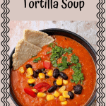 Creamy vegan tortilla soup in a black bowl garnished with black beans, corn, diced tomatoes, cilantro and tortilla chips.