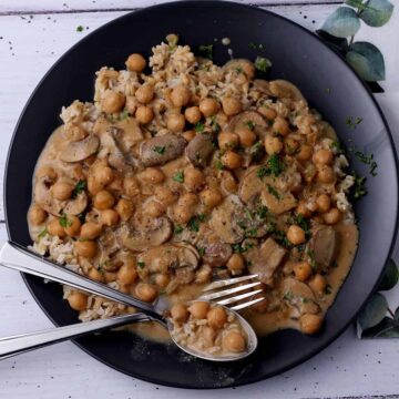 Creamed chickpeas and mushrooms over rice on a black plate with a spoon and fork.