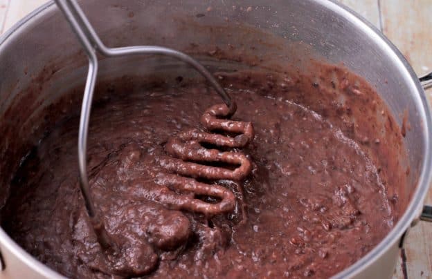 Black beans in a pan are mashed with a masher.