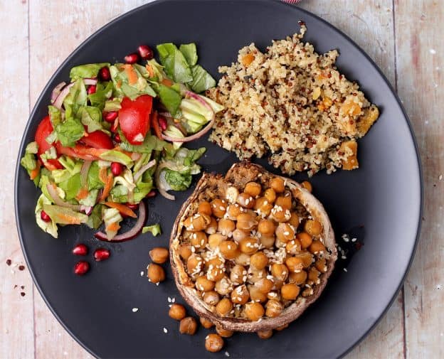 A black plate with a vegan stuffed Portobello mushroom with chickpeas, a green salad with pomegranate and quinoa.