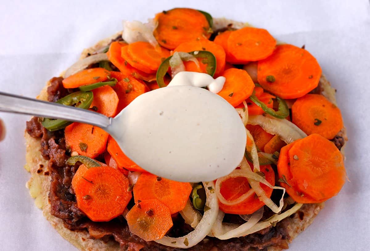 A spoonful of plant-based sour cream is held over a tostada with refried beans topped with pickled jalapenos, onions, and carrots.