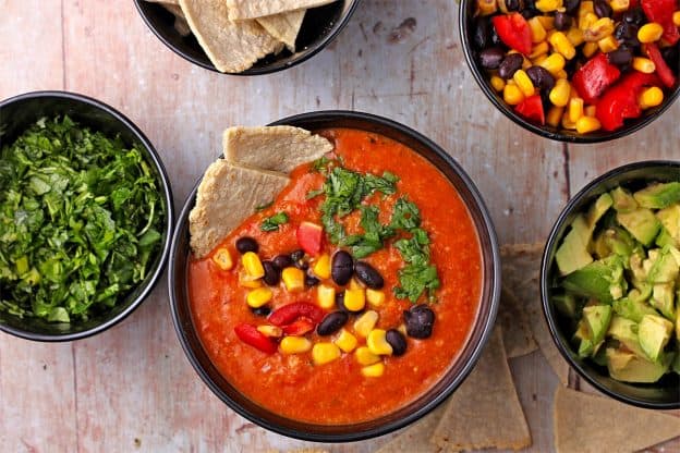 Tortilla soup in a black bowl with bowls of garnishes including diced avocado, cilantro, tortilla chips, and a bowl of mixed black beans, corn, and tomatoes.