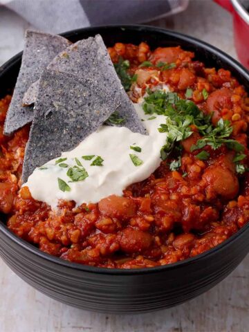 A black bowl of plant-based chili with plant-based sour cream, blue corn tortilla chips, and cilantro on top.
