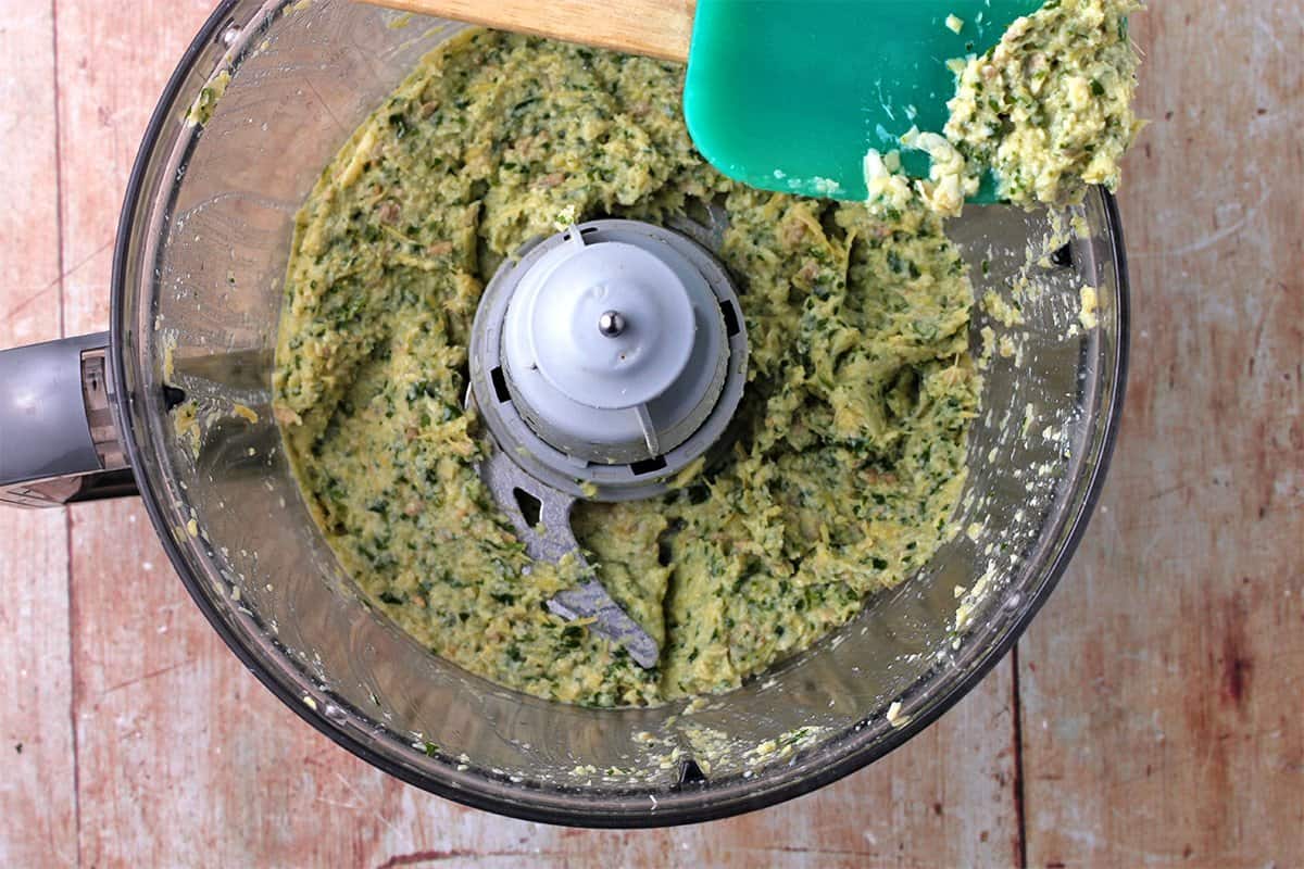 A food processor filled with artichoke pesto and a green scraper with pesto on the side.