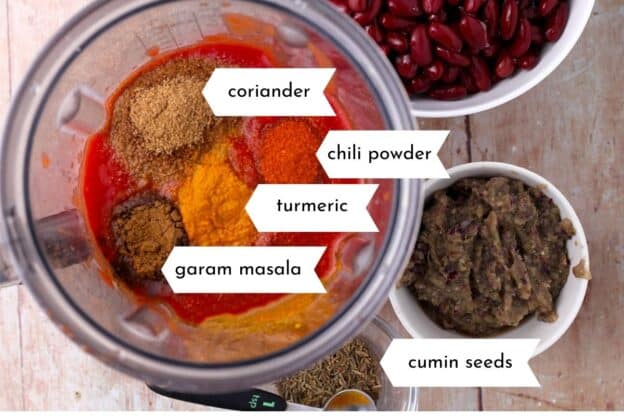 A blender is filled with tomatoes and spices that are labeled as cumin seeds, garam masala, turmeric, chili powder, and corinader.