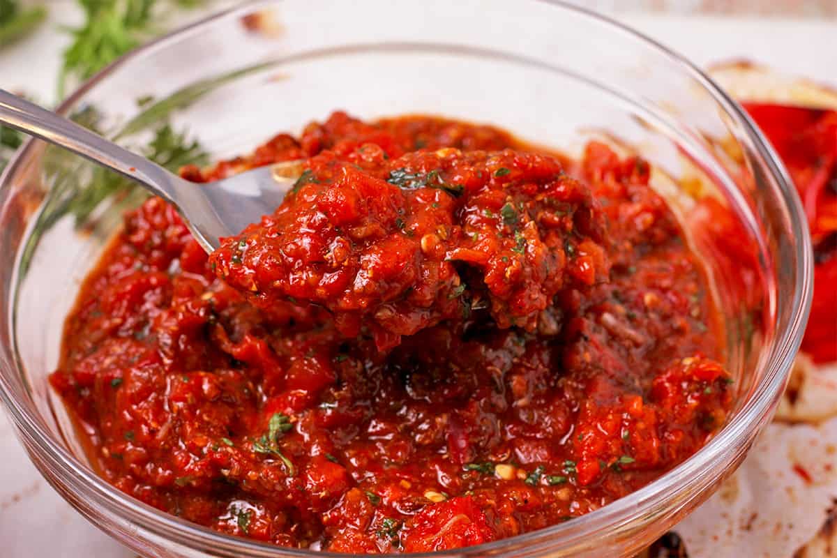 A spoon lifts red pepper harissa paste over a glass bowl.