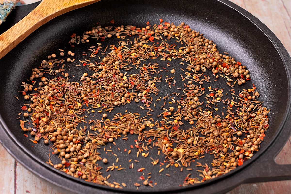 Coriander seeds, cumin seeds, caraway seeds, and red chili flakes in a black skillet.