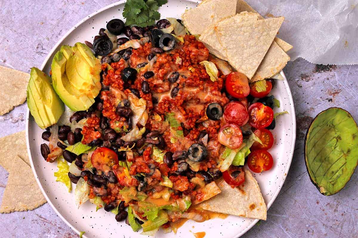 A taco salad on a white plate with chipotle dressing, bulgur taco meat, black olives, lettuce, tomatoes, avocados, tortilla chips, and dressing.
