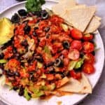 A salad with lettuce, tomatoes, black beans, black olives, bulgur wheat taco meat, tortilla chips, avocados, and dressing.