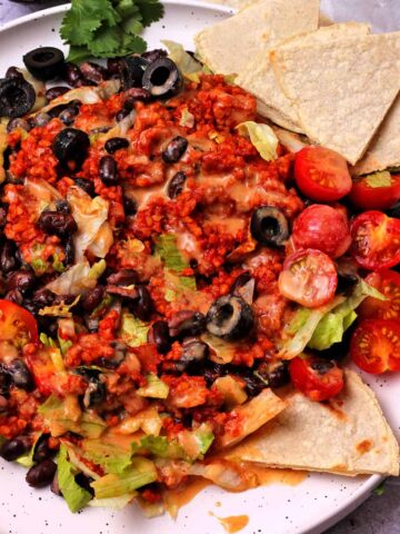 A salad with lettuce, tomatoes, black beans, black olives, bulgur wheat taco meat, tortilla chips, avocados, and dressing.