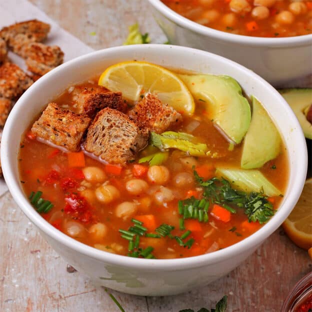 A bowl of chickpea harissa soup with bread cubes, avocado, lemon wedges, celery leaves, and chopped chives.