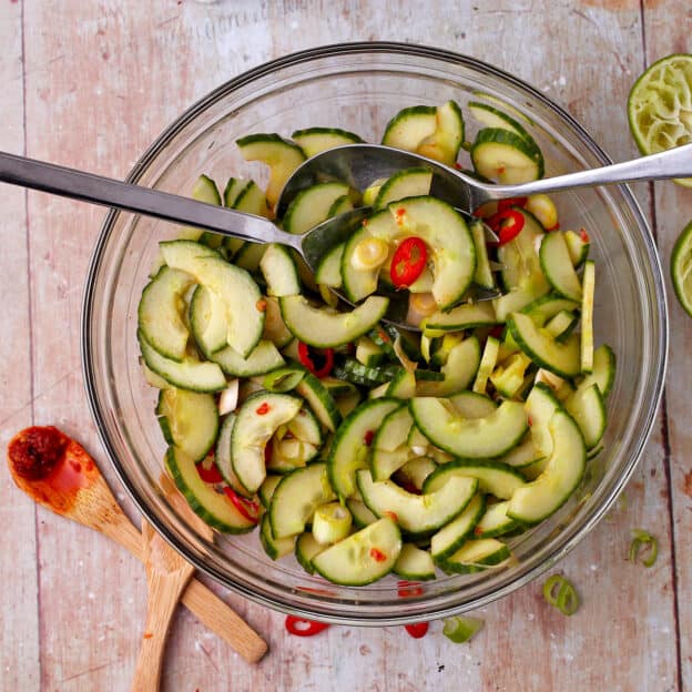 A glass bowl of cucumber salad with red chili, sliced scallions and a fork and spoon.