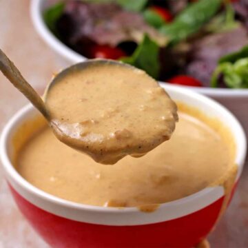 A ladle lifts mustard tahini dressing and it runs over the side of a red bowl.