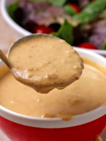 A ladle lifts mustard tahini dressing and it runs over the side of a red bowl.