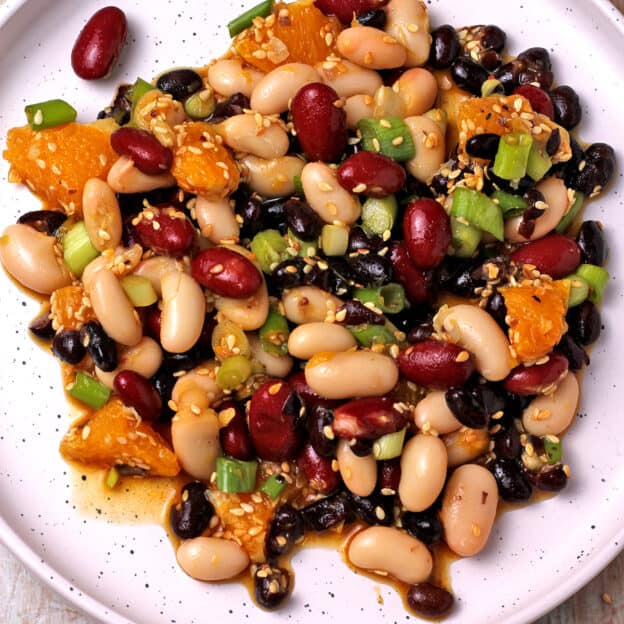 A white plate with white, black, and red beans, sesame seeds, orange segments, and sliced scallions.