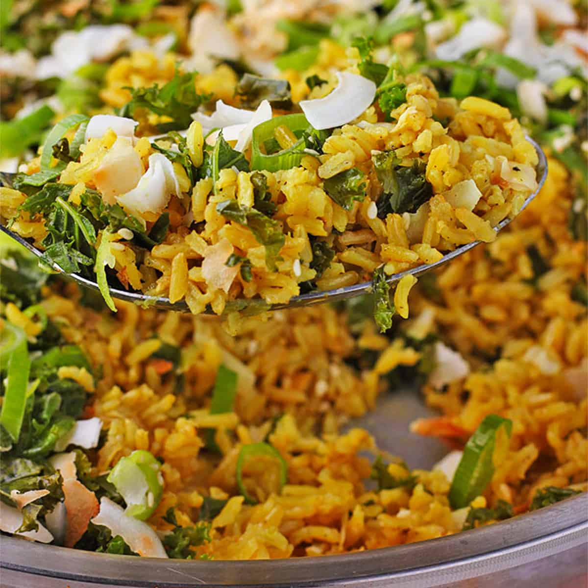 A spoonful of turmeric rice with scallions, coconut flakes, sesame seeds, and scallions is lifted over a skillet of rice.