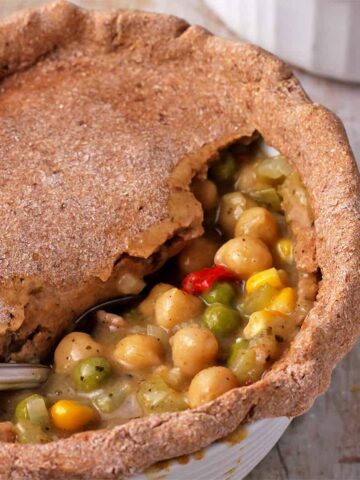 A spoon is dipped in a vegan pot pie with chickpeas, vegetables, and gravy topped with a whole wheat crust.