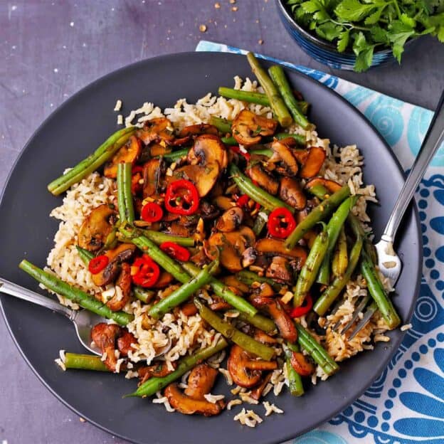 Green bean mushroom stir fry with red chili over rice on a black plate.