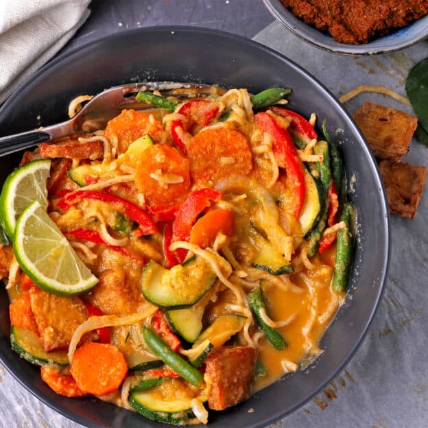 A bowl is filled with vegetable mango curry with noodles and baked tofu.
