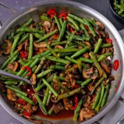 A skillet with green beans, mushrooms, sliced red chilis, coriander seeds, and cilantro.