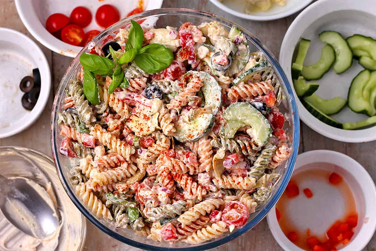 Creamy Italian pasta salad in a glass bowl with bowls of ingredients.
