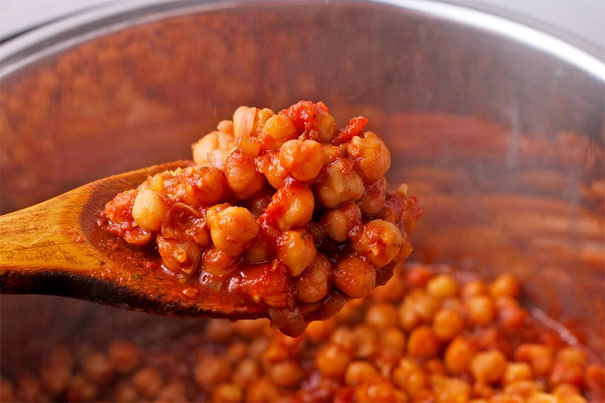 A wooden spoon filled with chickpeas in Kapama sauce is held over the Instant Pot.