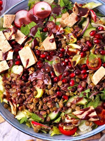 A bowl of salad with lentils, tomatoes, cucumber, radish, pita chips, pomegranate, and dressing.