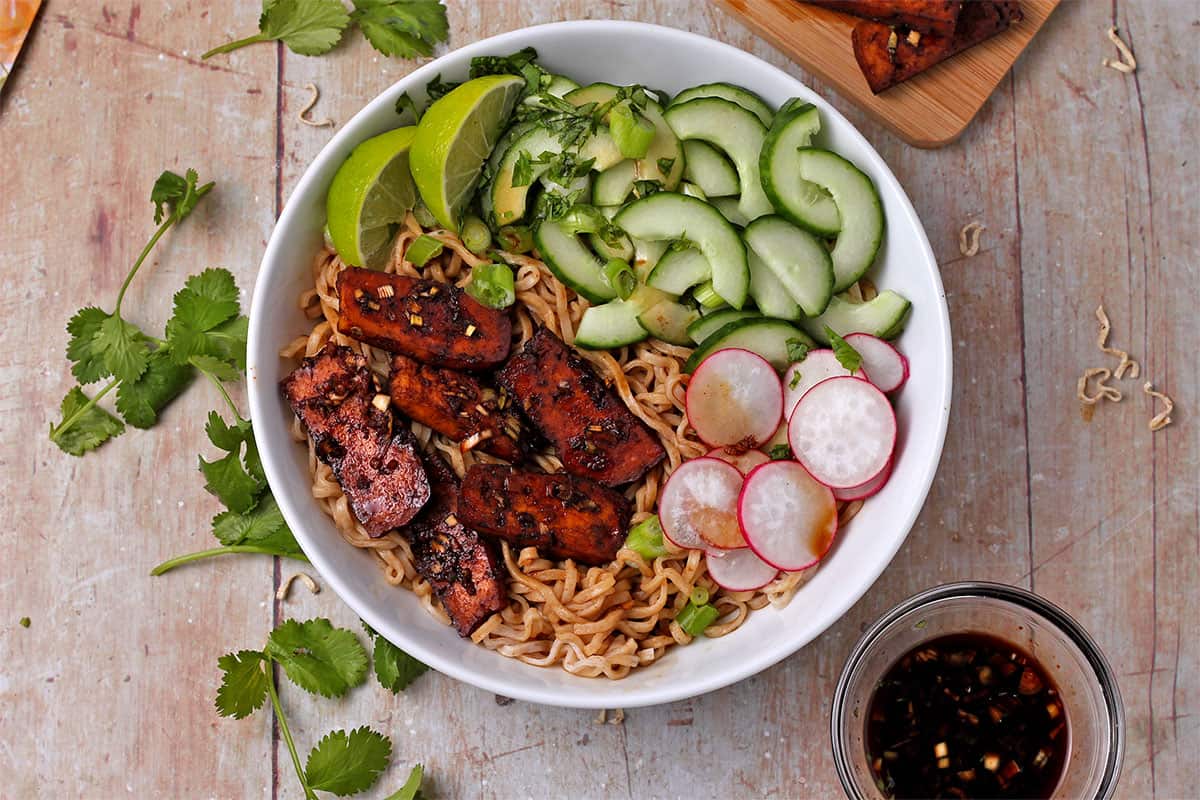Overhead of a bowl with baked tofu, noodles, cucumbers, radishes, cilantro, and scallions.