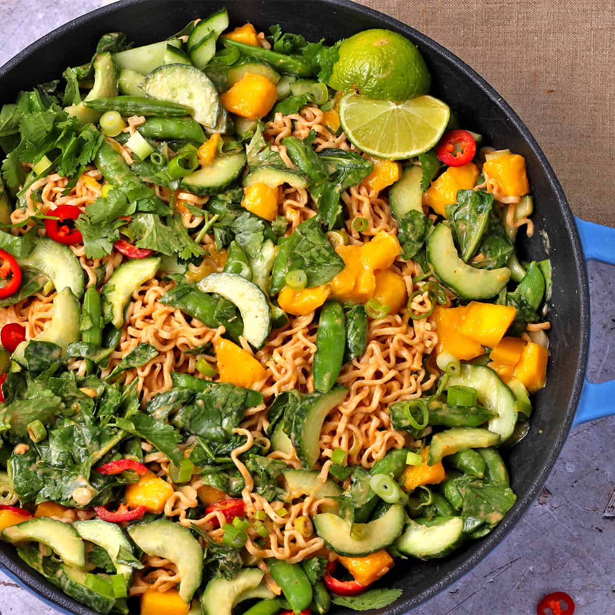 A skillet with a salad with cucumber, scallions, snap peas, mango, red chili, noodles, and peanut sauce.