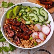 A bowl with baked tofu, noodles, lime wedges, cucumbers, radishes, scallions, and cilantro with a bowl of dressing.