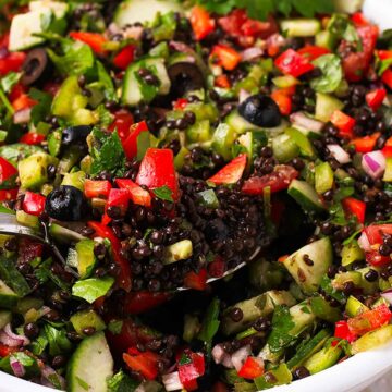A spoon is filled with cooked black lentils and Greek salad and held over a bowl.