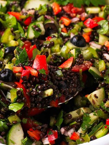 A spoon is filled with cooked black lentils and Greek salad and held over a bowl.