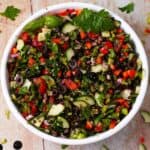 A bowl filled with cucumbers, tomatoes, olives, peppers, herbs, and black lentils.
