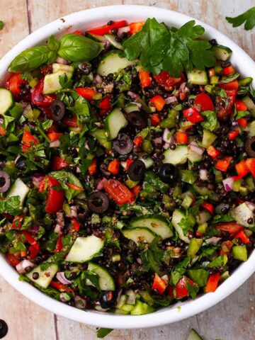 A bowl filled with cucumbers, tomatoes, olives, peppers, herbs, and black lentils.