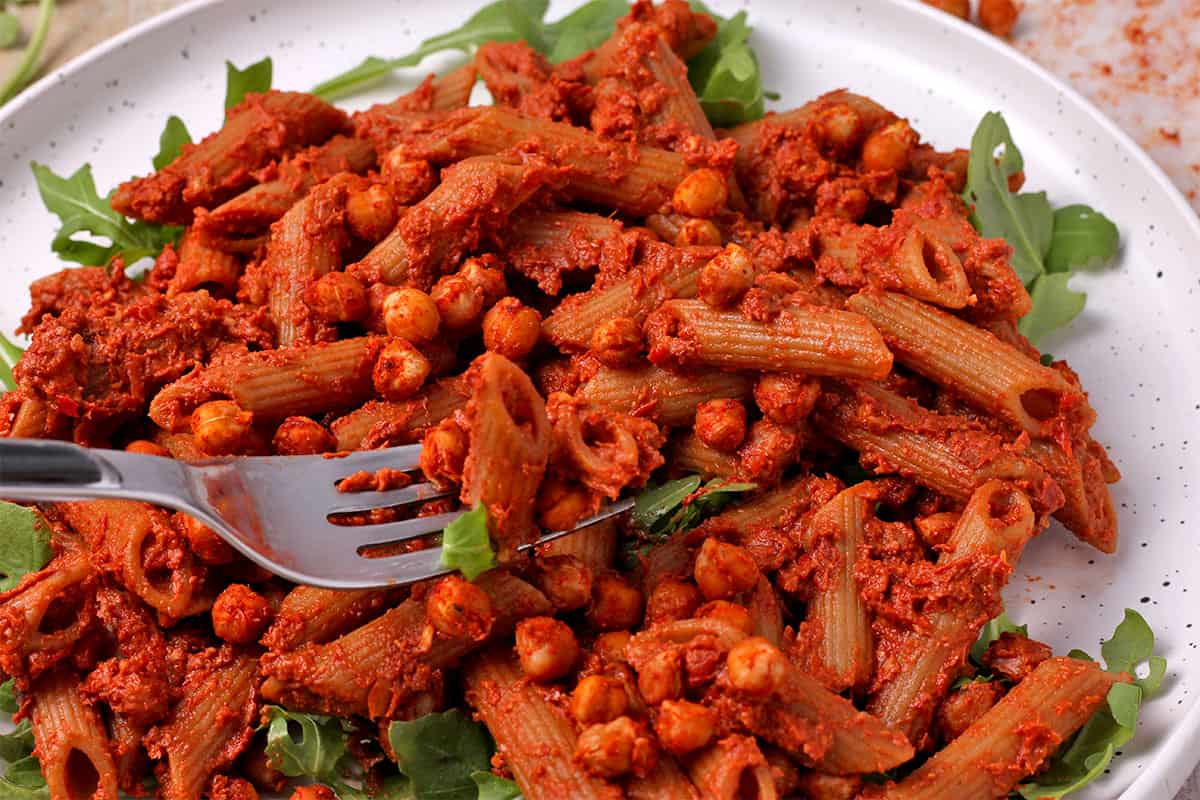 A fork lifts penne pasta with red pesto and seasoned chickpeas.