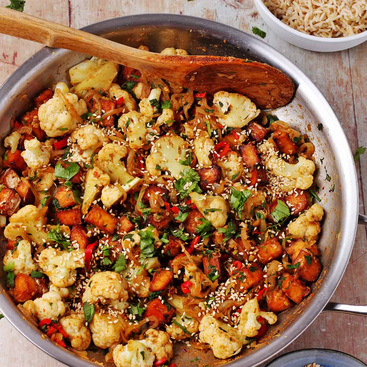 A skillet filled with stir fried cauliflower and tofu with onions, red chili, cilantro, and sesame seeds.