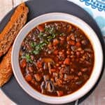 A bowl of mushroom barley soup with carrots, celery, and onion on a black plate with bread on the side.