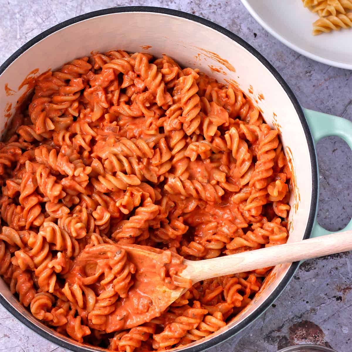 A blue pot filled with pasta with pink sauce is stirred with a spoon.