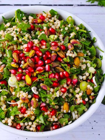 A white bowl filled with pearl couscous, pomegranate, herbs, and pistachios.