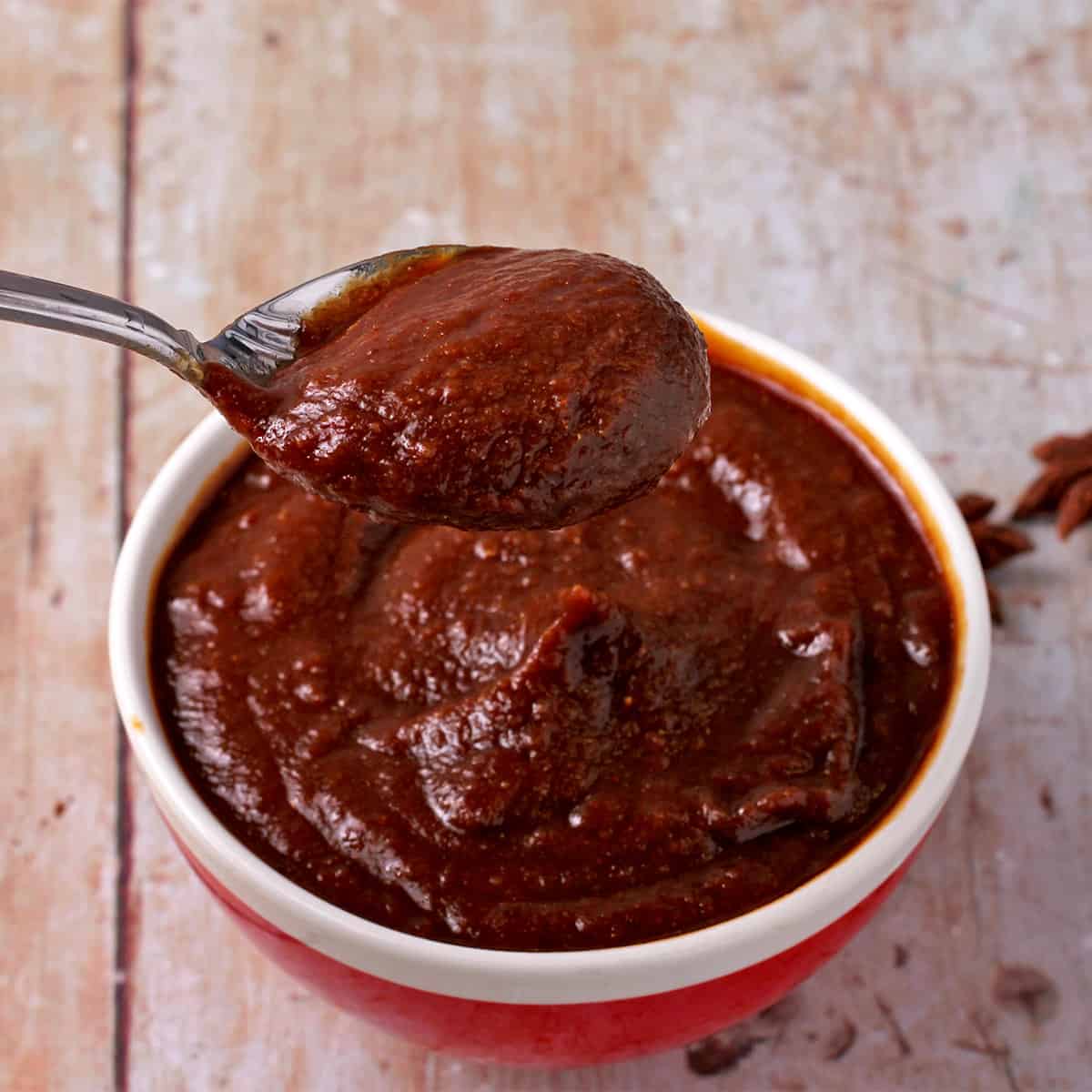 A spoonful of homemade hoisin sauce is lifted over a red bowl filled with sauce.