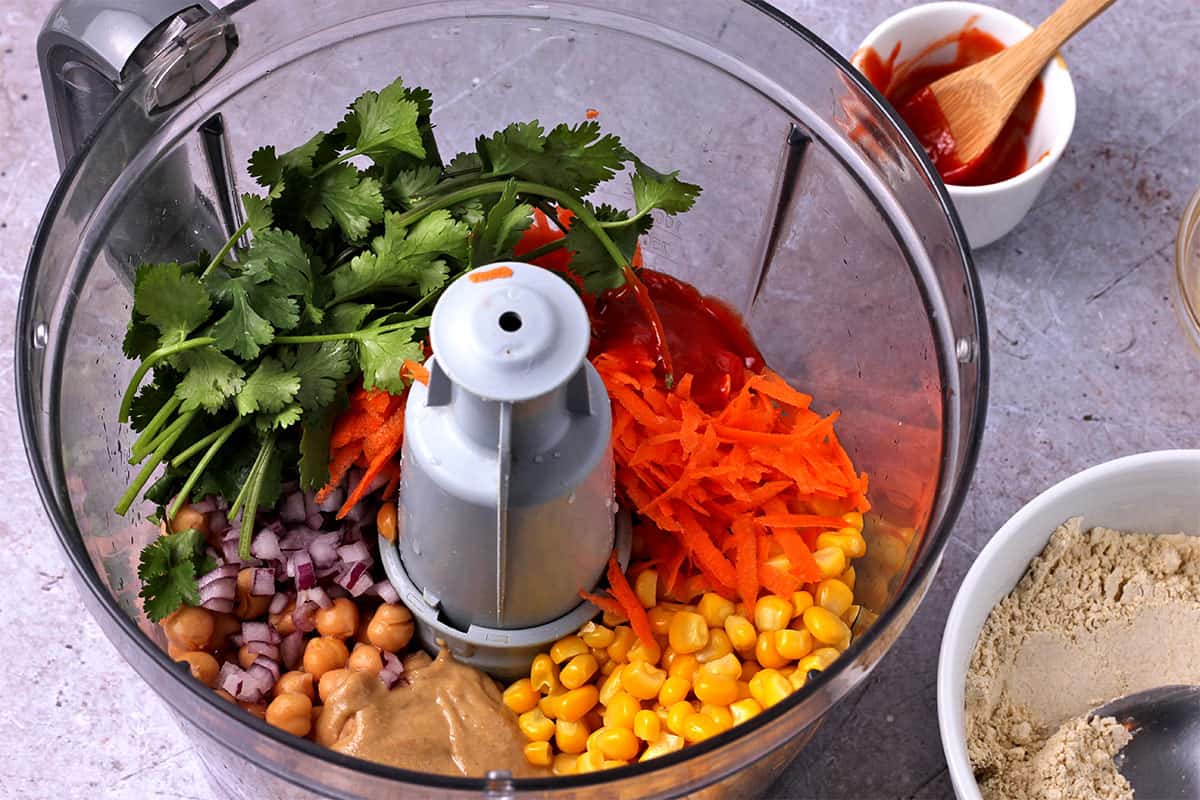 Corn, chickpeas, tahini, red onions, carrots, cilantro, and sriracha are placed in a food processor.