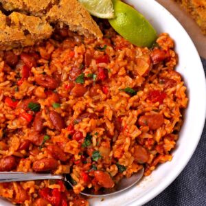 Cooked pinto beans and rice with tomatoes and red peppers are in a white bowl with a silver spoon, 2 cornbread slices, and lime wedges.