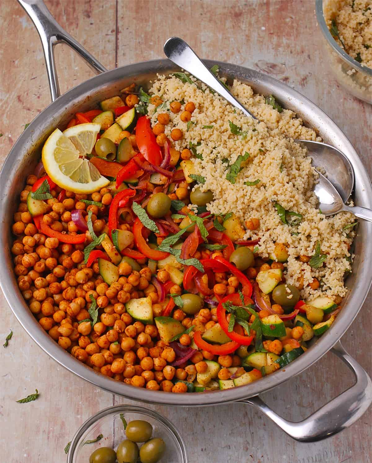 A sliver skillet filled with chickpea and couscous salad with green olives and fresh mint.